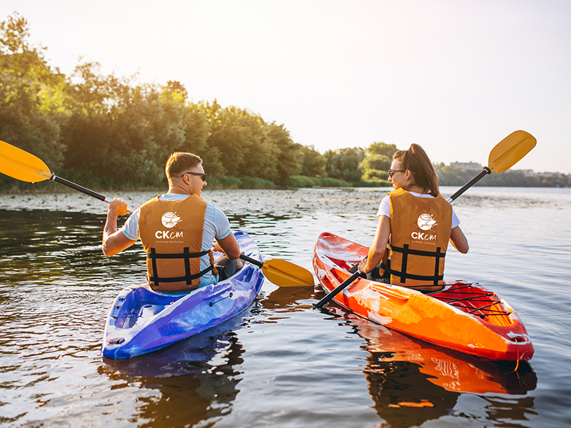 Photographie d'un homme et d'une femme de dos naviguant en kayak sur une rivière calme en pleine nature lors du coucher du soleil.